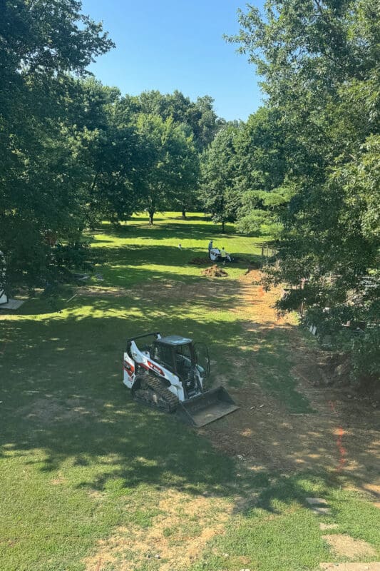 Skid steer parked in the backyard at the start of excavation for the sidewalk, walking trail, with orange marking paint and stakes laying out the route across the lawn