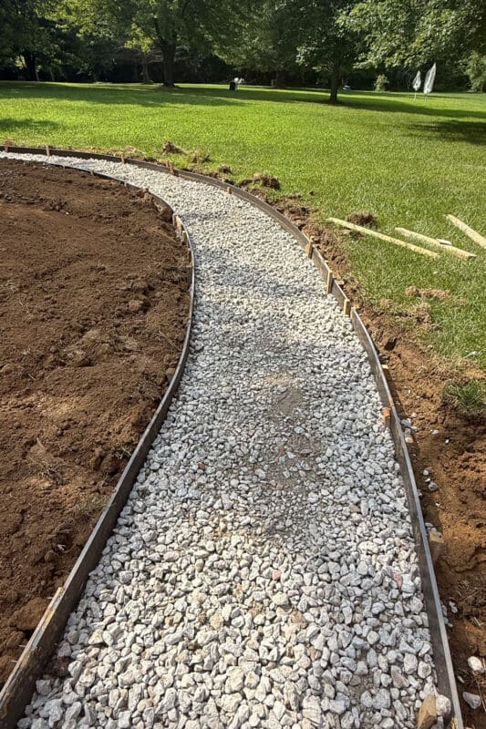 Gravel base spread and leveled between wooden forms along the curving route of the concrete path in the backyard, with fresh dirt on one side and lawn on the other
