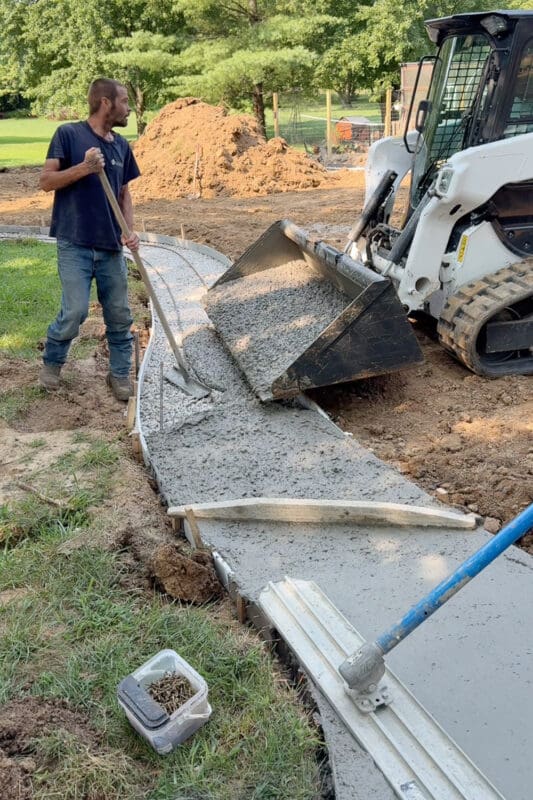 Skid steer bucket dumping wet concrete into the forms for the concrete path in the backyard while a worker spreads it with a come-along rake