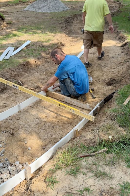 Worker kneeling to set flexible white forming material along the edge of the paved sidewalk in the backyard while checking it with a long level