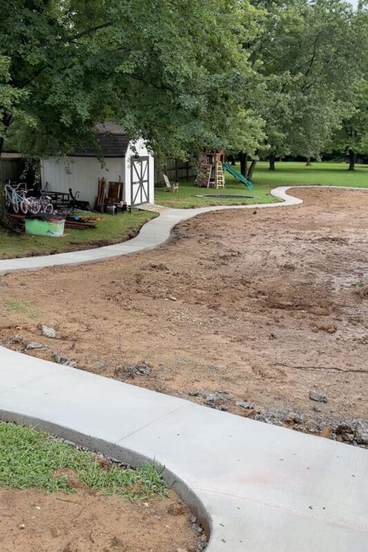 Concrete path in the backyard curving past a white shed with kids bikes parked alongside it, leading toward a playset and in ground trampoline