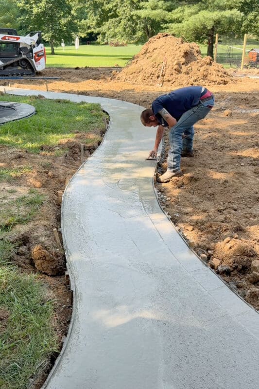 Worker bending over to trowel the edge of the freshly poured concrete path in the backyard, with the smooth wet surface curving away toward the trampoline and lawn