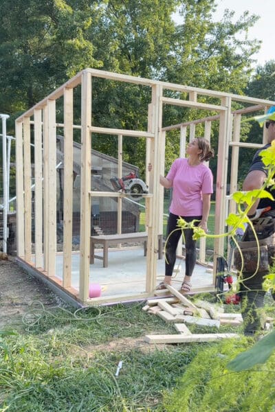 Woman standing inside a framed in outbuilding with all four 2x4 walls framed up on the concrete slab, looking up at the top plate with the old coop visible behind