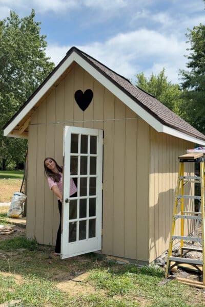 Woman peeking around the open glass paned door of a finished DIY chicken coop with a heart cutout in the gable