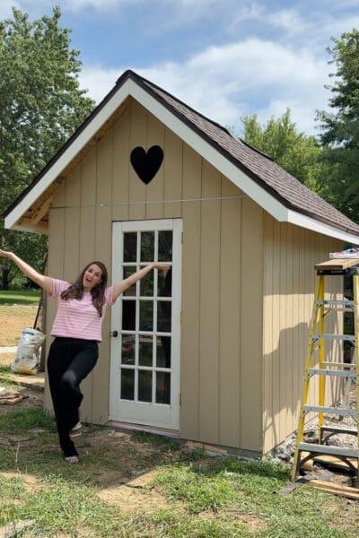 Tan board and batten chicken coop with a brown shingled peaked roof, white trim, and a heart shaped vent, with ladders still set up alongside from the build