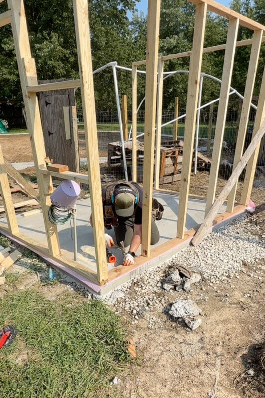 Framed walls standing on a concrete slab with a pink sill plate gasket visible along the bottom plate, and a man kneeling inside fastening the frame down