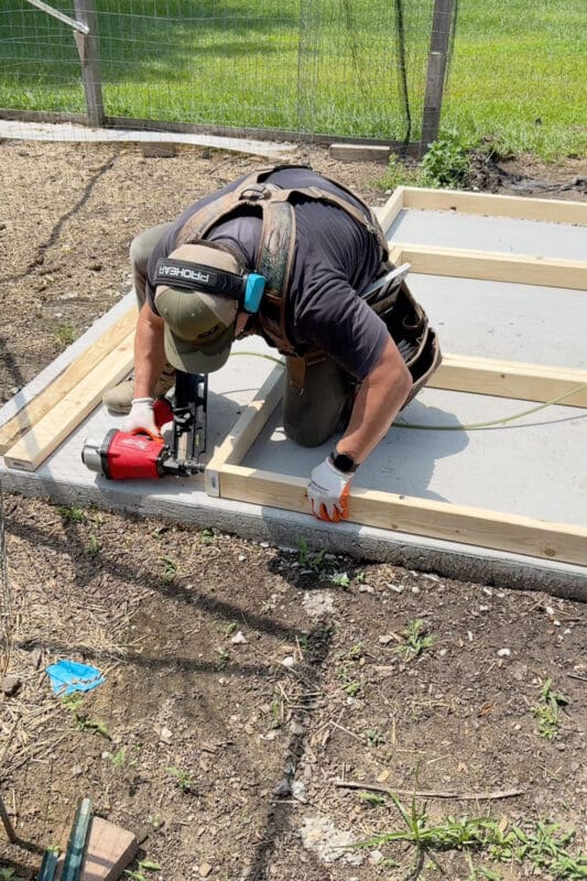 Man using a framing nailer to assemble lumber wall plates directly on the concrete slab at the start of a backyard project