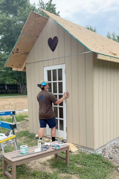 Man holding a white glass paned door in place in the framed opening of a chicken coop with the heart cutout above and bare roof sheathing overhead