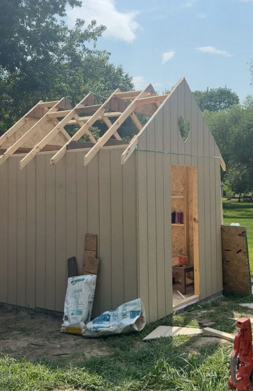 Chicken coop with tan siding installed showing a heart shaped cutout in the gable, a framed door opening, and rafter tails cut to length for the eaves