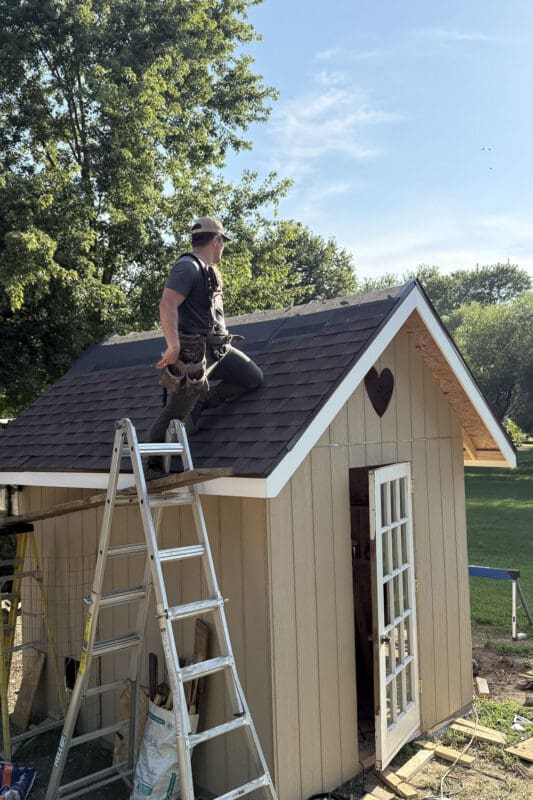 Man kneeling on a ladder jack platform installing brown architectural shingles on the peaked roof of a chicken coop with the glass paned door open below
