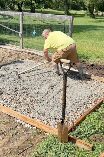Worker in a yellow shirt smoothing wet concrete with a rake inside a wooden form set over gravel for a n outbuilding foundation