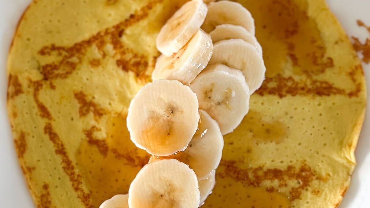 A high-protein pancake on a plate, topped with a line of banana slices and drizzled with golden maple syrup, capturing a close-up of the delicious details.
