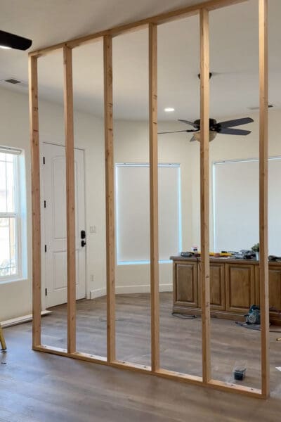 Freestanding stud frame for a DIY room divider wall, secured at the ceiling and floor, with the office area, windows, and wooden desk visible through the open studs.