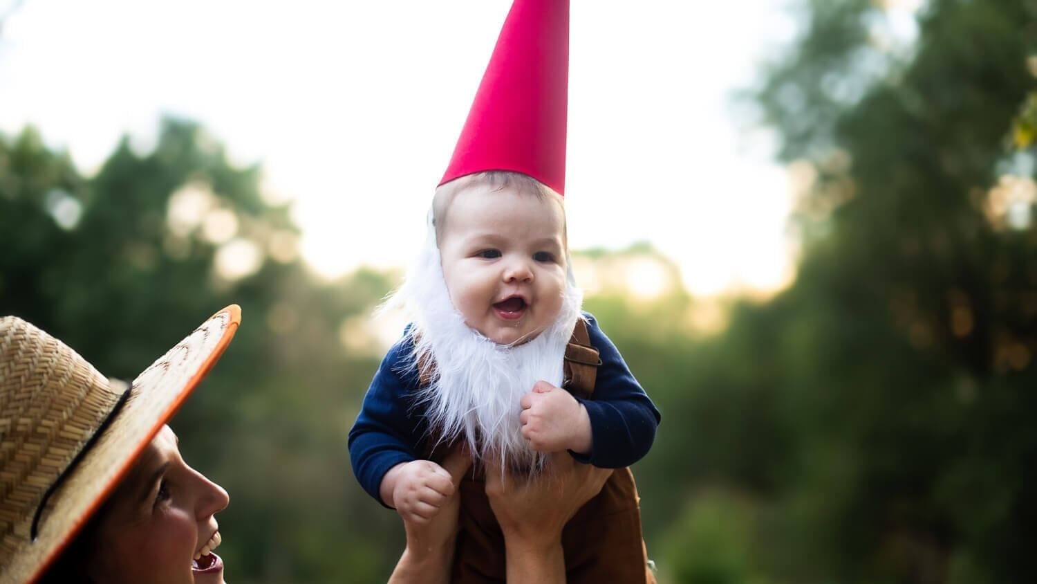 Baby dressed in a handmade garden gnome costume with a red hat, white beard, and brown overalls being lifted up outdoors by a smiling mom wearing a straw gardener hat.