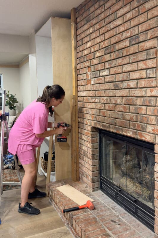 Crystel securing a shorter 2x4 to the brick at mantel height, stacked against the full-height board on the DIY fireplace built ins