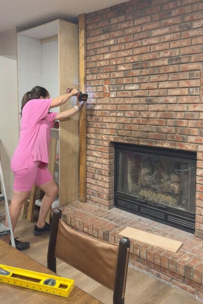 Crystel drilling a full-height 2x4 into the brick beside the plywood cabinet box 