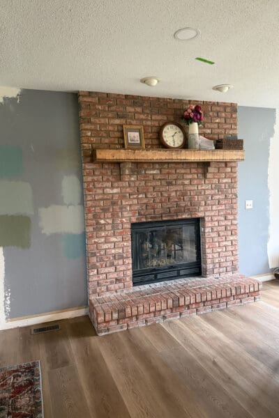 The brick fireplace before the DIY fireplace built ins, with a rustic wood mantel and bare walls on either side marked with gray and green paint swatches