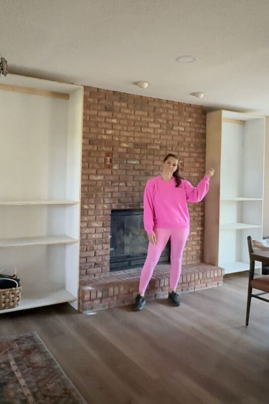 Crystel standing at the brick fireplace with both raw plywood cabinet boxes set in place on either side, before trim was added to the DIY fireplace built-ins