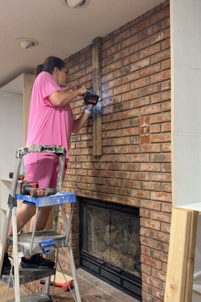 Crystel on a step ladder attaching a vertical 2x4 to the center of the brick chimney above mantel height for the DIY fireplace built ins