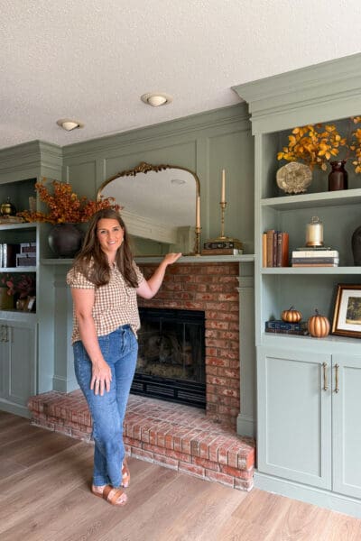 Crystel standing beside the finished DIY fireplace built ins, gesturing toward the mantel with sage green shelves and cabinets flanking the brick fireplace