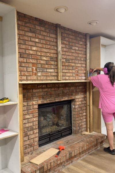 Crystel attaching the first mantel board across the 2x4 blocking on the brick fireplace, spanning between the two plywood cabinet boxes on the DIY fireplace built ins
