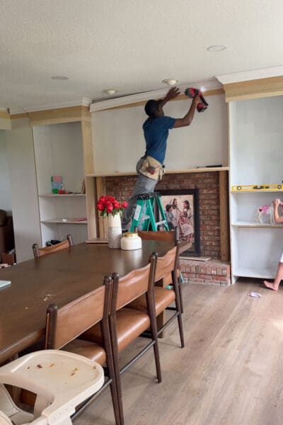 A hired carpenter on a ladder nailing crown molding along the ceiling above the DIY fireplace built ins