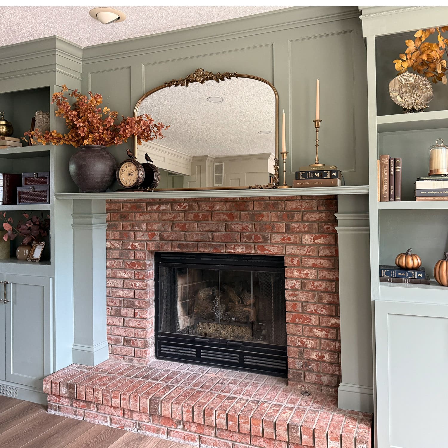 Sage green DIY fireplace built ins framing the original brick fireplace, with an arched gold mirror above the mantel and shelves styled with brass candlesticks and fall stems