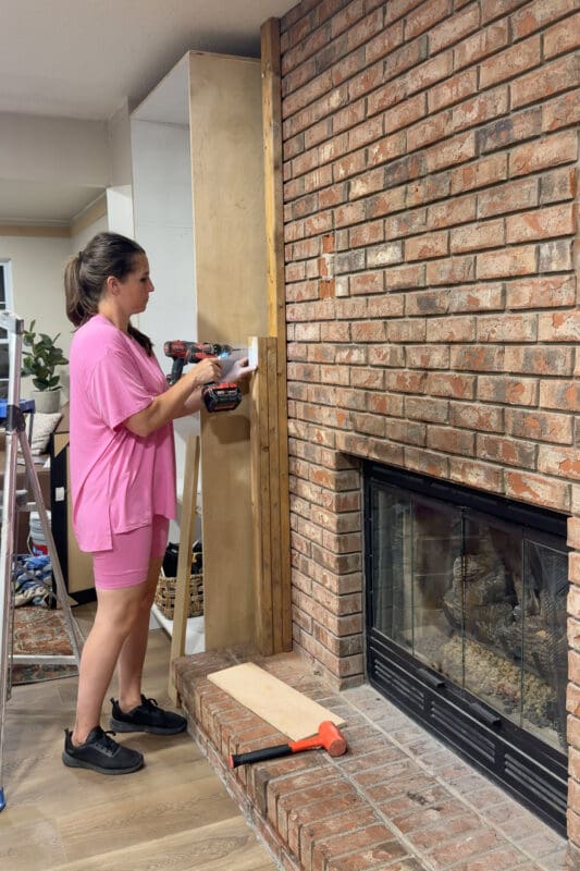 Crystel holding a 2x4 in place against the brick at mantel height, building out the blocking that will carry the trim on the DIY fireplace built ins