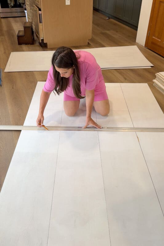 Crystel drawing shelf lines with a pencil and straightedge across primed plywood panels laid out on the floor for the DIY shelves she is making