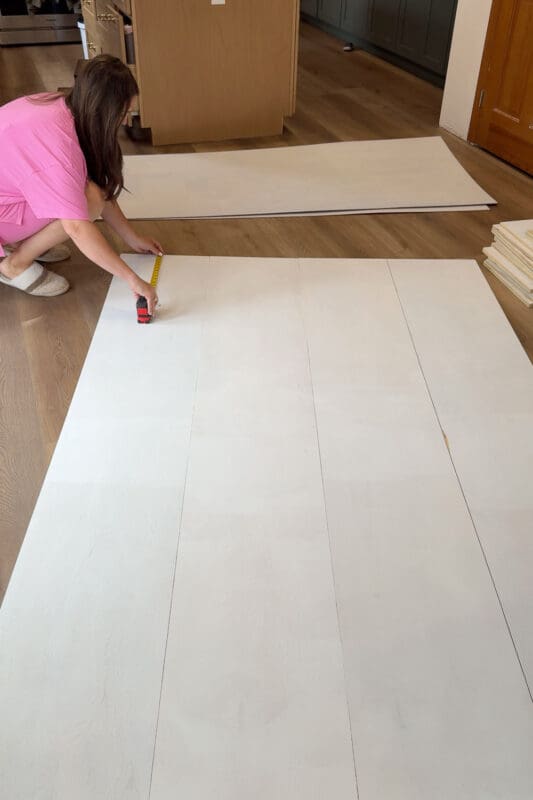 Crystel kneeling with a tape measure to mark shelf placement on a primed plywood panel before assembling DIY cabinets for her dining room.