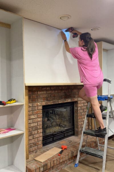 Crystel attaching a large primed plywood panel to the wall above the mantel, covering the brick chimney on the DIY fireplace built ins