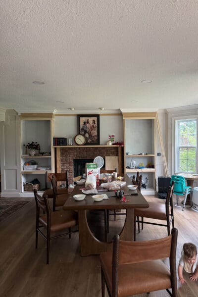 The DIY fireplace built ins sitting unfinished in raw plywood with crown molding installed, holding books and everyday items in the lived-in dining room