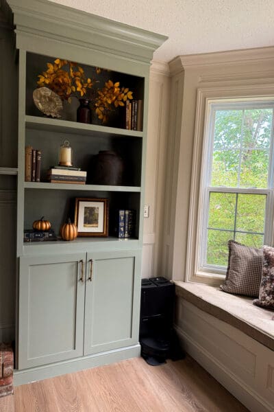 The right side of the DIY fireplace surround with styled shelves, brass pulls, and shaker doors, next to the window bench in the corner of the room