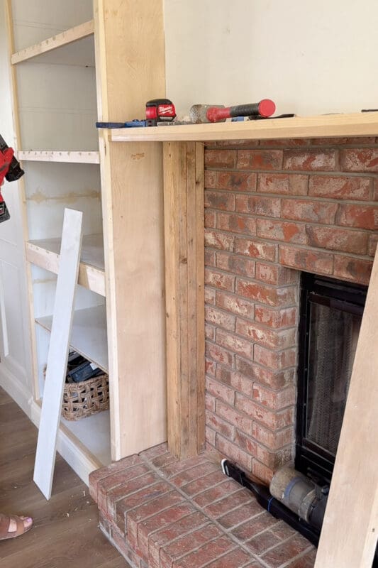 The 2x4 column beside the firebox partly wrapped in plywood, with the next piece leaning ready against the DIY fireplace surround shelves