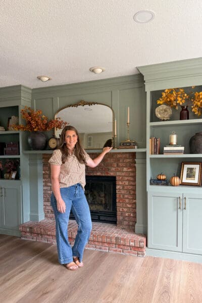 Crystel standing beside the finished DIY fireplace surround, with the sage green mantel and column trim framing the original brick