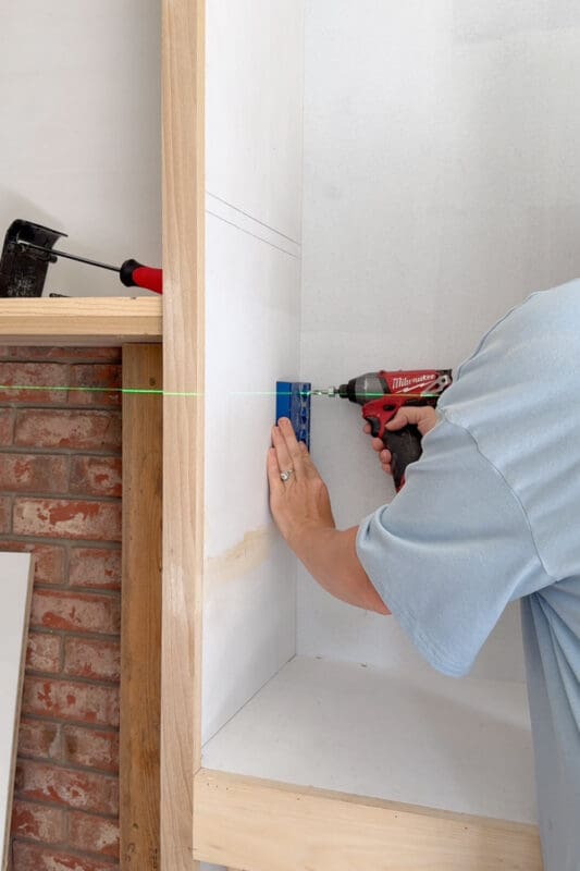 Crystel drilling shelf pin holes with a Kreg jig held against the laser line inside the DIY fireplace surround cabinet