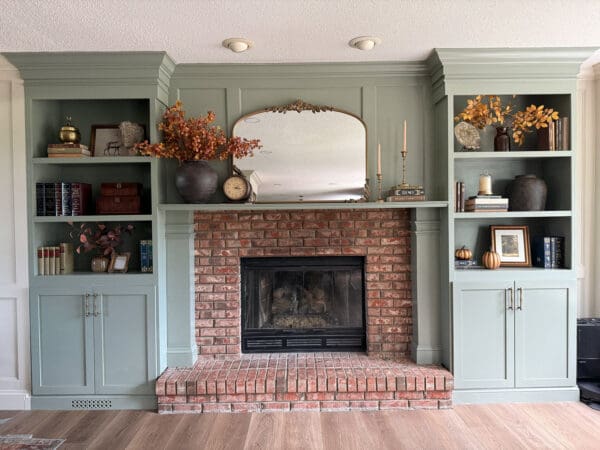 The finished DIY fireplace surround in sage green, with styled shelves, brass pulls, and an arched gold mirror above the trimmed mantel and brick firebox
