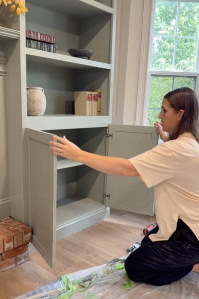 Crystel kneeling to hang a sage green shaker door on the lower cabinet of the DIY fireplace surround, with books and pottery already styled on the shelves above