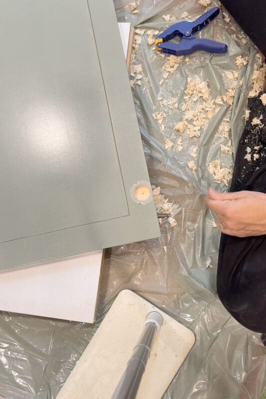 A freshly bored hinge cup hole in the back of a sage green cabinet door, surrounded by wood shavings on the drop cloth for the DIY fireplace surround