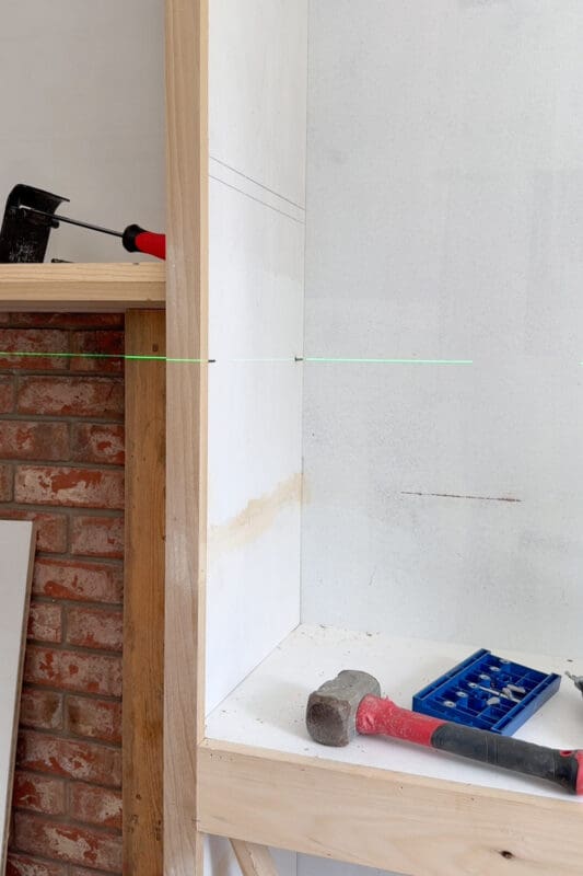 A green laser line marking shelf pin placement across the inside of the cabinet, with a Kreg shelf pin jig and mallet resting on the shelf below
