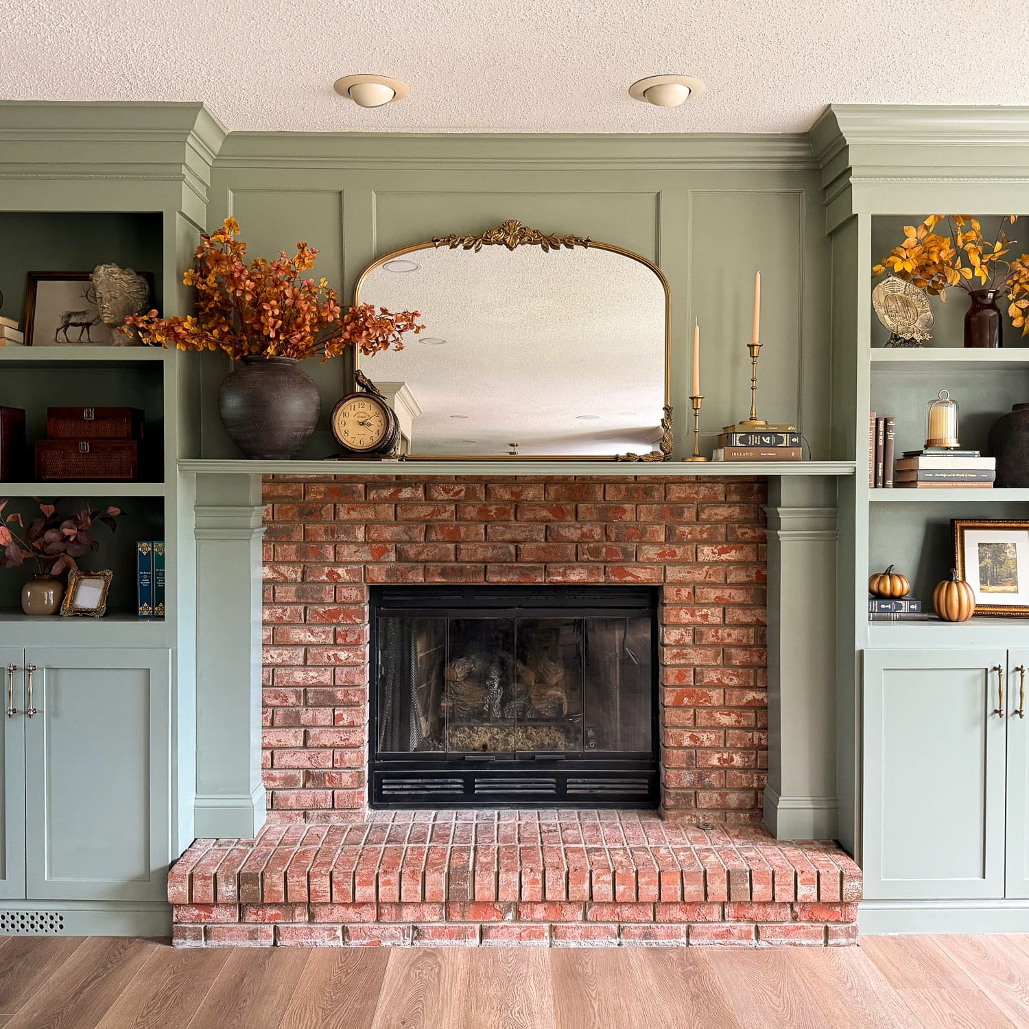 The completed DIY fireplace surround in sage green, with paneled columns on either side of the brick firebox and a trimmed mantel below the gold mirror
