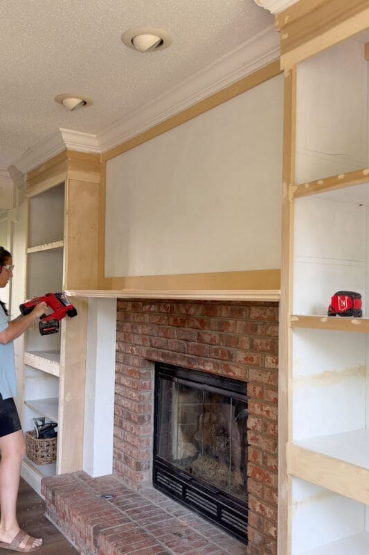 Crystel nailing decorative trim along the front edge of the doubled plywood mantel on the DIY fireplace surround