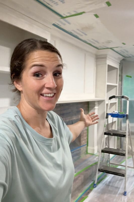 Crystel smiling in front of the DIY fireplace surround with the brick, ceiling, and hearth masked off in plastic and tape, primed and ready for paint
