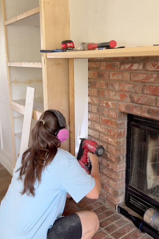 Crystel nailing a plywood panel onto the 2x4 column beside the brick firebox to build out the DIY fireplace surround
