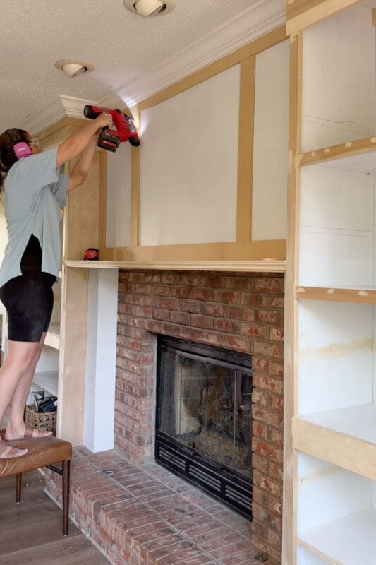 Crystel on a chair nailing trim strips onto the panel above the mantel to create the framed overmantel detail on the DIY fireplace surround