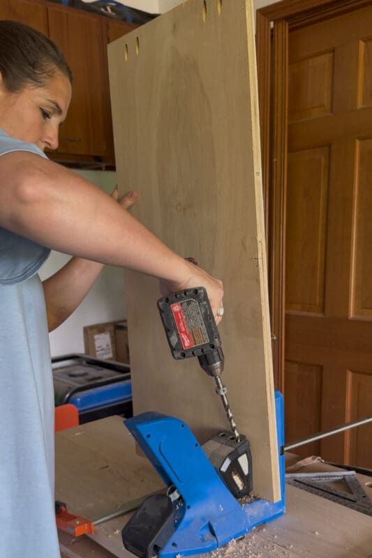 Crystel drilling pocket holes along the edge of a plywood shelf with a Kreg jig.