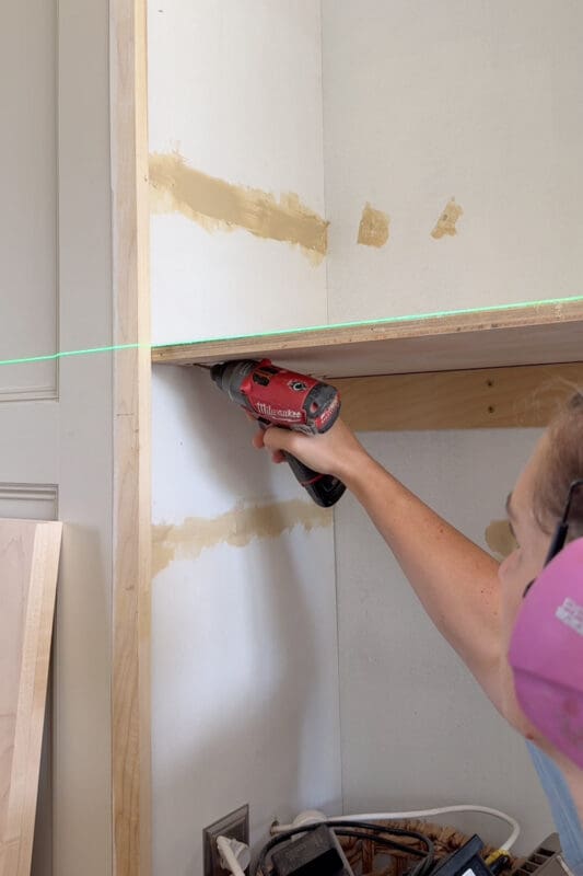 Crystel driving pocket screws to attach the relocated shelf inside the cabinet, with a green laser line marking the new height