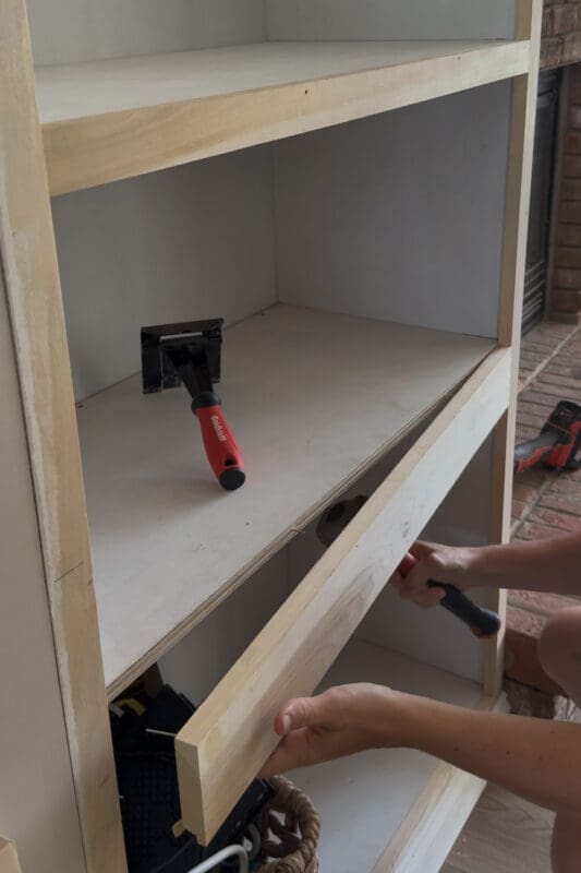 Prying a face frame board loose from the plywood shelf on the of a built-in cabinet, using a pry bar to reach the fasteners underneath