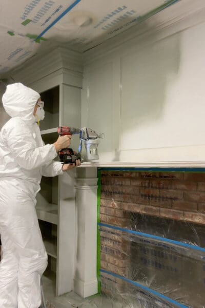 Crystel in a protective suit and respirator spraying sage green paint onto the DIY fireplace surround columns and overmantel panel