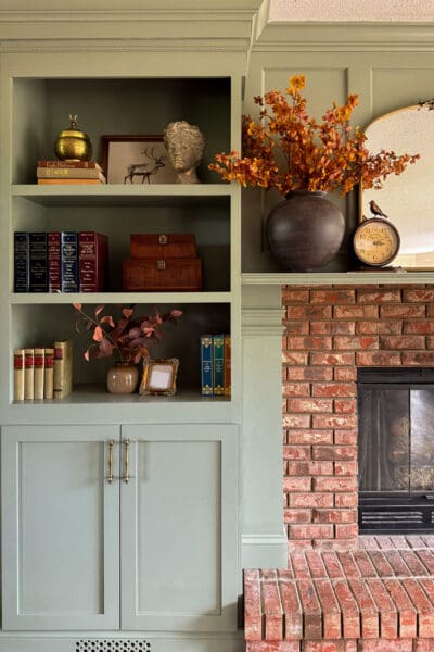 Close view of the styled sage green shelves beside the brick fireplace, with vintage books, a brass jar, and a dark vase of fall stems on the DIY fireplace surround
